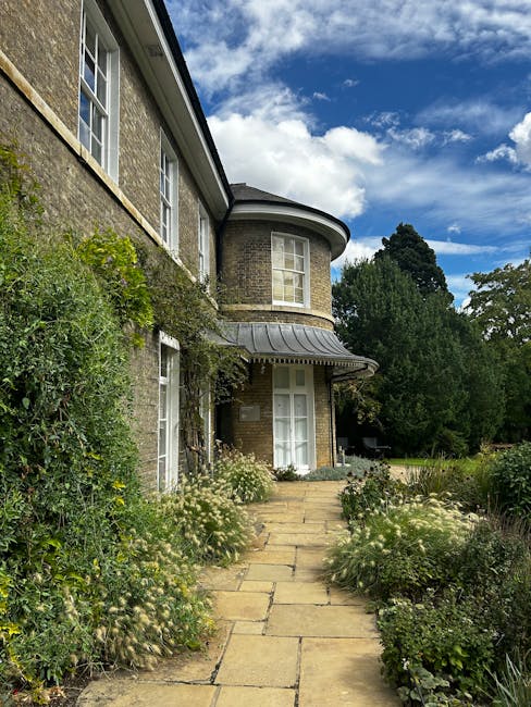 A traditional brick residential building with white-framed windows and a curved bay window on the upper floor, set against a partly cloudy sky. The garden features a stone pathway leading to the entra